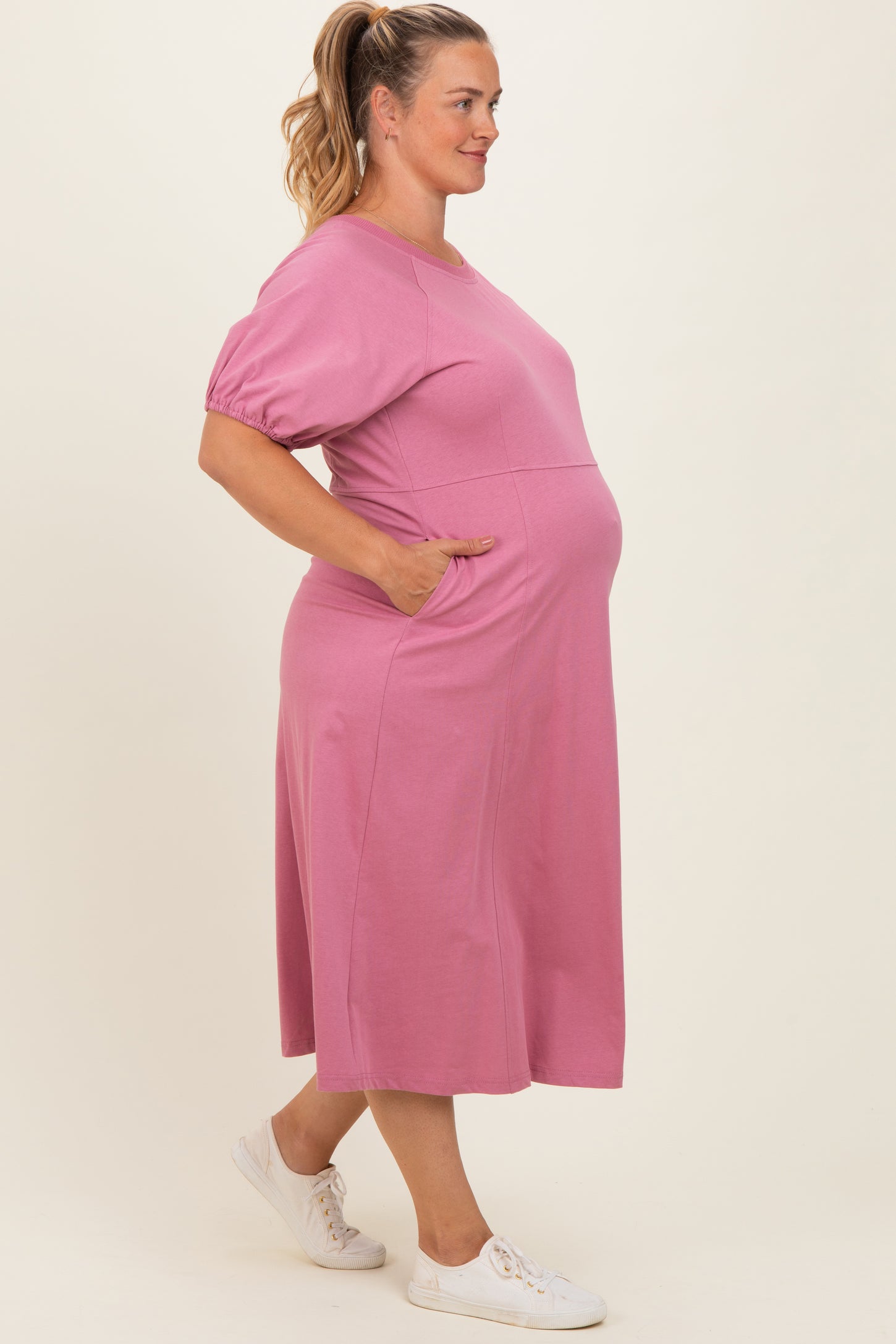 woman wearing a pink maternity plus midi dress with short sleeves and pockets, showing her baby bump
