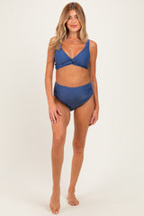 woman wearing a blue maternity bikini with a twist front top and high-waisted bottoms, standing barefoot on a neutral background