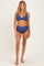 woman wearing a blue maternity bikini with a twist front top and high-waisted bottoms, standing barefoot on a neutral background