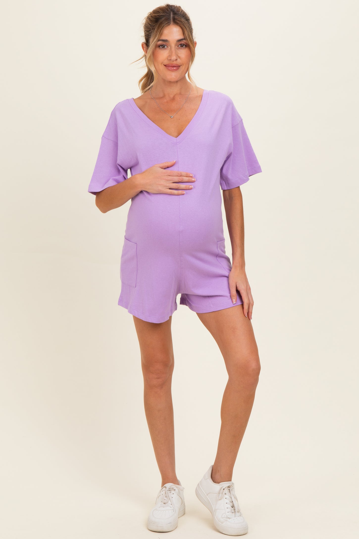 woman wearing a lavender maternity romper with short sleeves and pockets, holding her baby bump