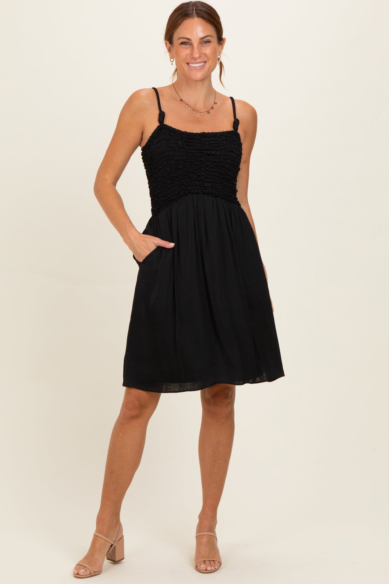 woman wearing a black smocked sundress with adjustable straps and pockets, standing against a neutral background