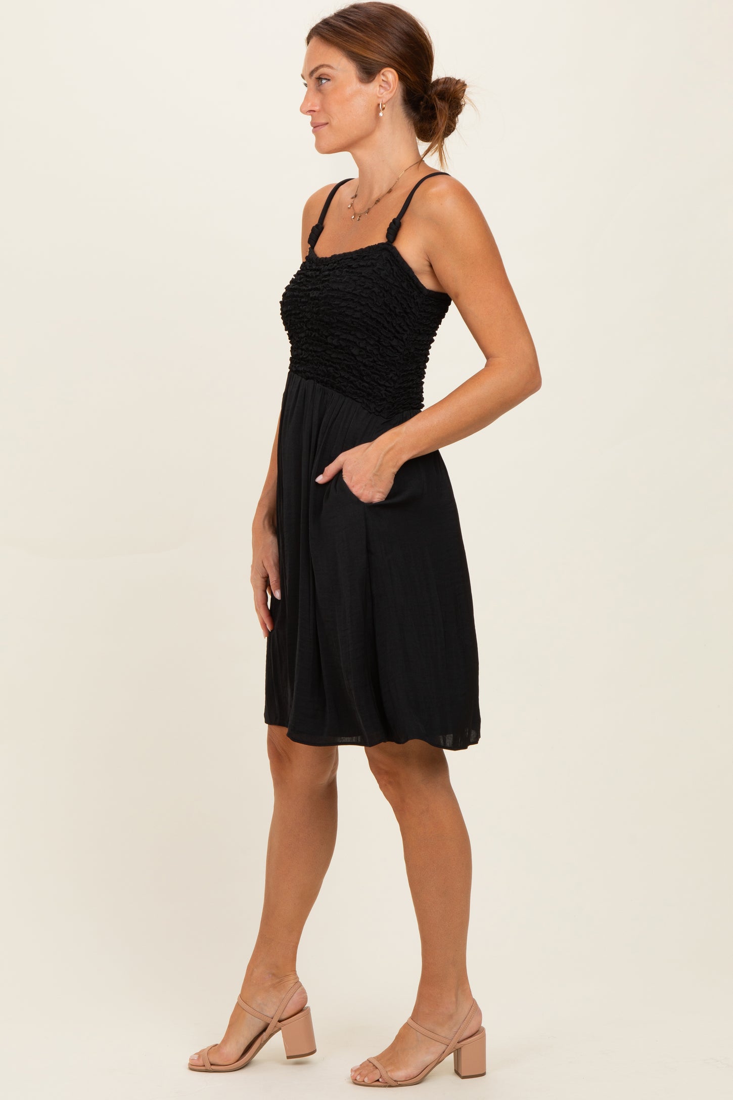 woman wearing a black smocked sundress with adjustable straps and side pockets, paired with beige block heel sandals