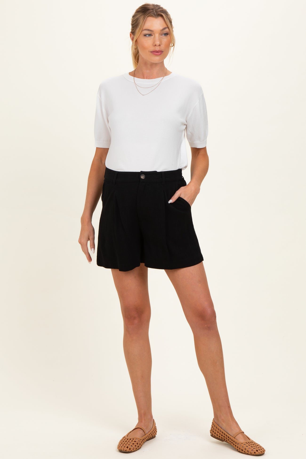 woman wearing a white short sleeve top and black maternity shorts, standing with hands in pockets