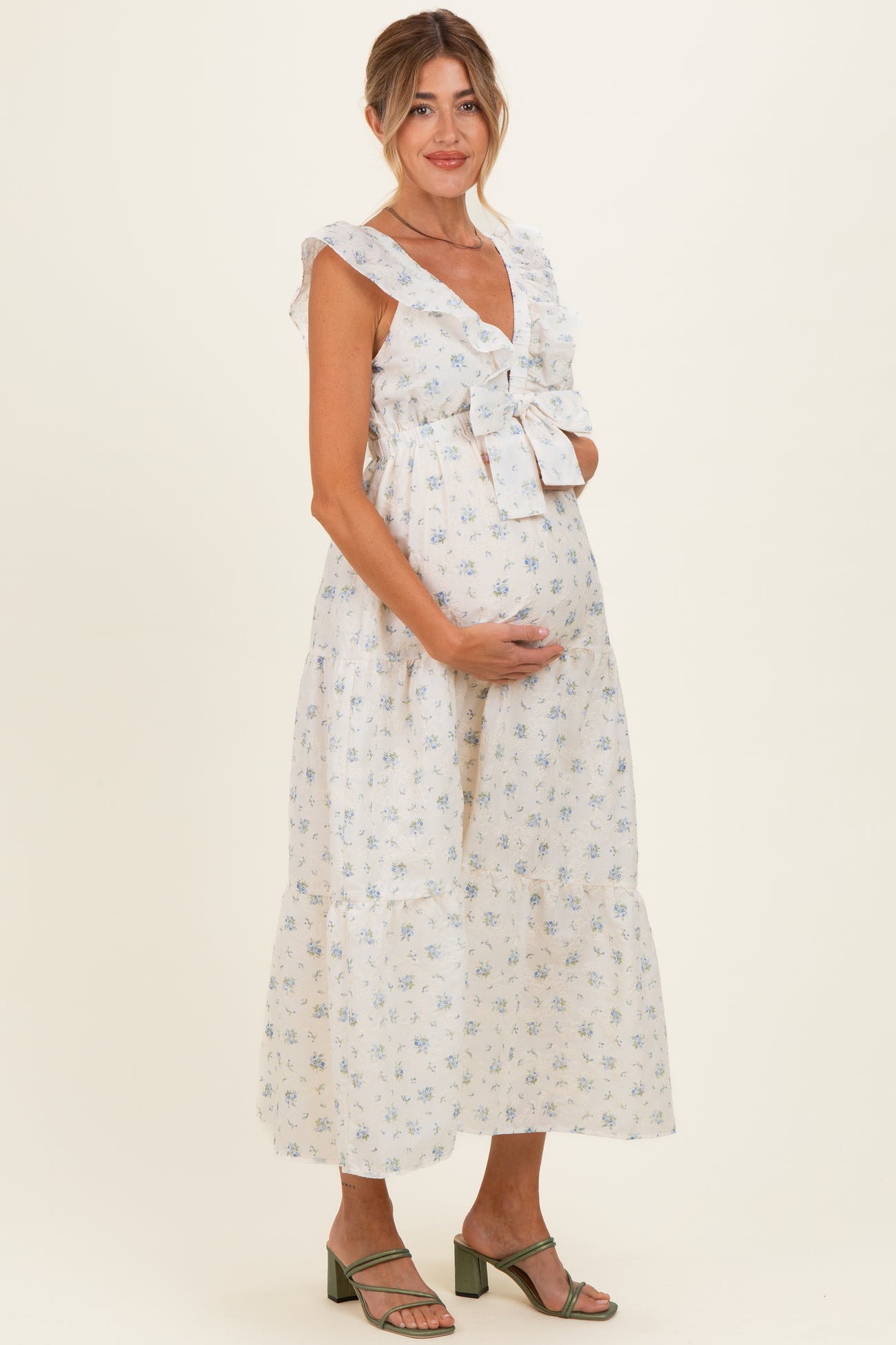 woman wearing a white floral maternity dress with ruffled sleeves and a tie detail, holding her baby bump