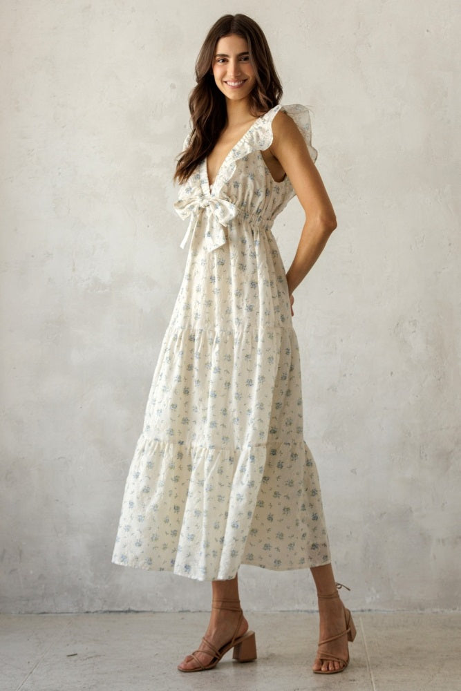woman wearing a white floral maxi dress with ruffled straps and a bow waist detail, standing against a textured background