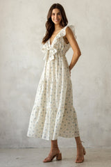 woman wearing a white floral maxi dress with ruffled straps and a bow waist detail, standing against a textured background