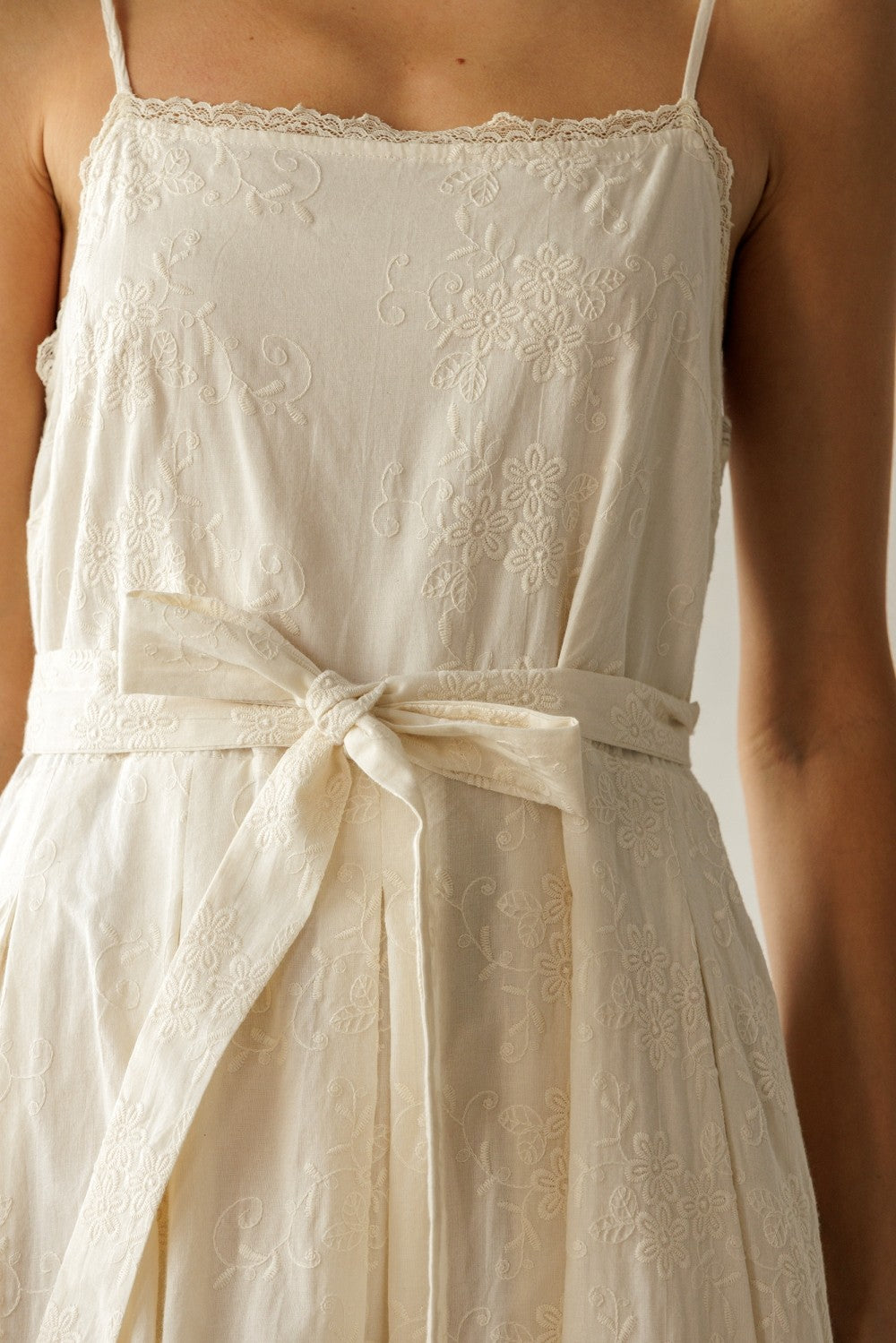 woman wearing a cream sleeveless dress with floral embroidery and a tied waist detail