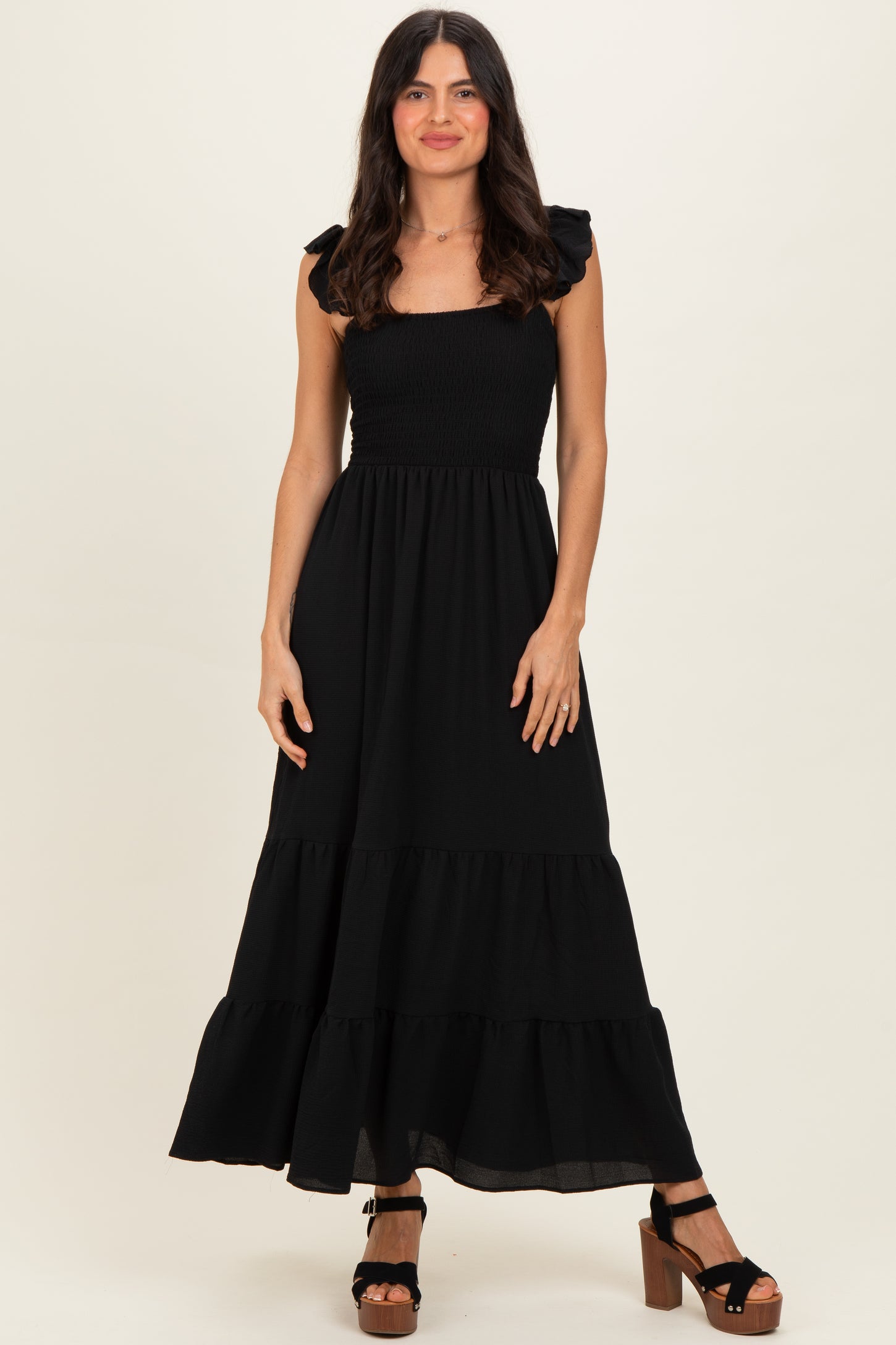 woman wearing a black maxi dress with ruffled sleeves and a tiered hem, standing confidently on a neutral background