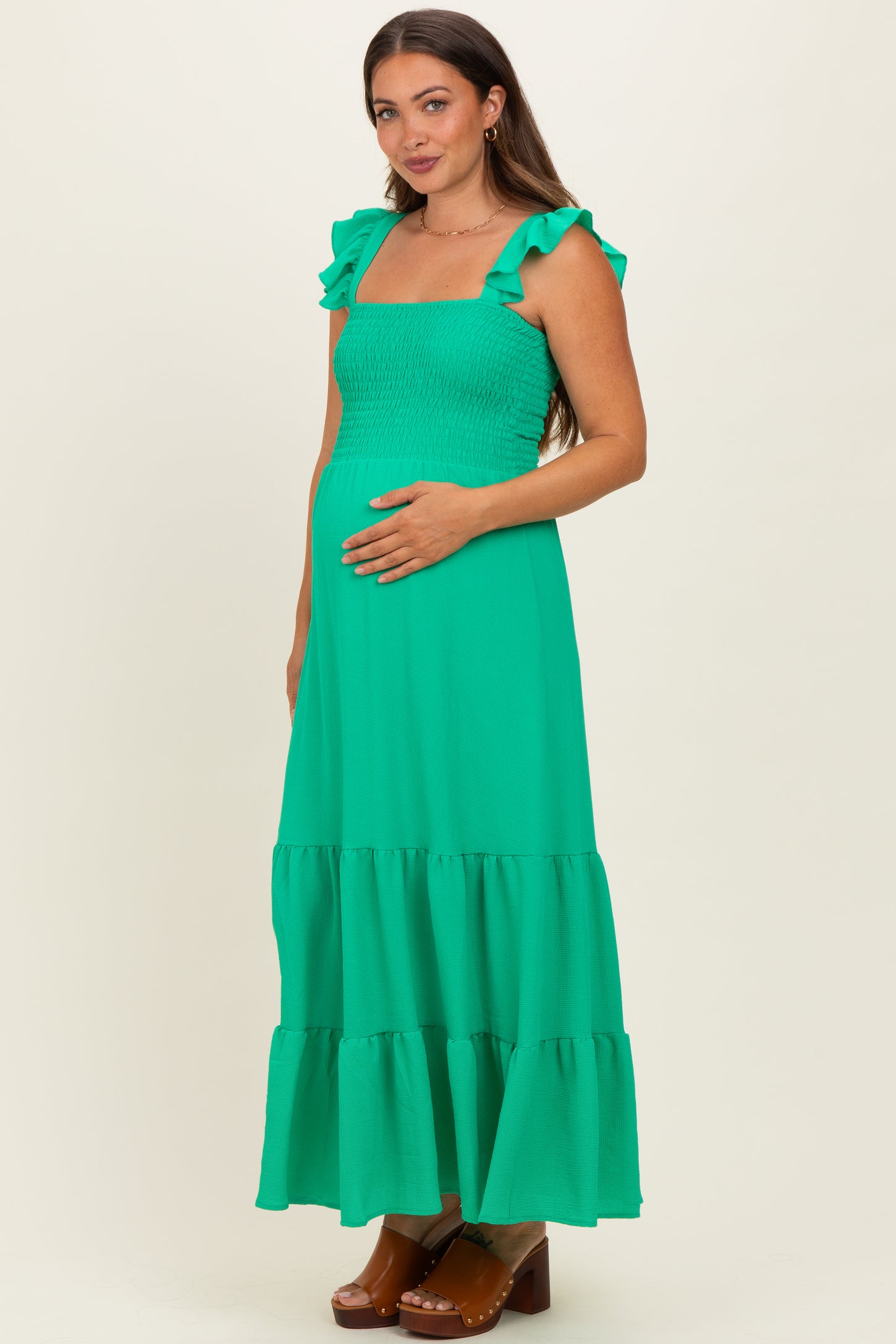 woman wearing a green maternity maxi dress with ruffle straps and a smocked bodice, holding her baby bump