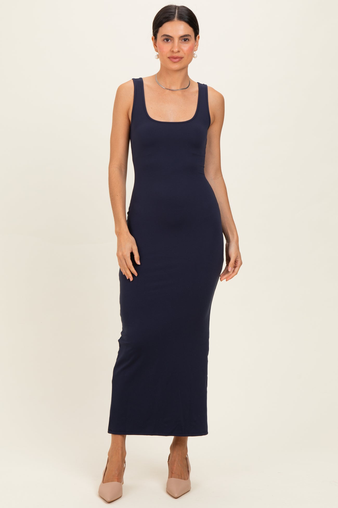 woman wearing a navy blue sleeveless maxi dress, posing with a neutral background and nude high heels