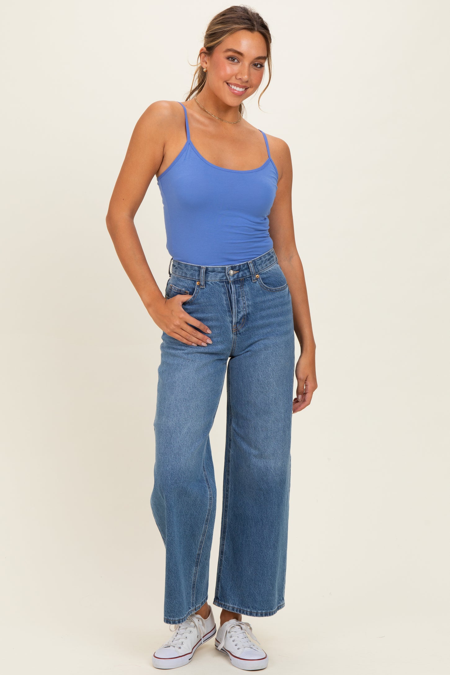 woman wearing a light blue tank top and high-waisted wide-leg jeans, posing with one hand on her hip