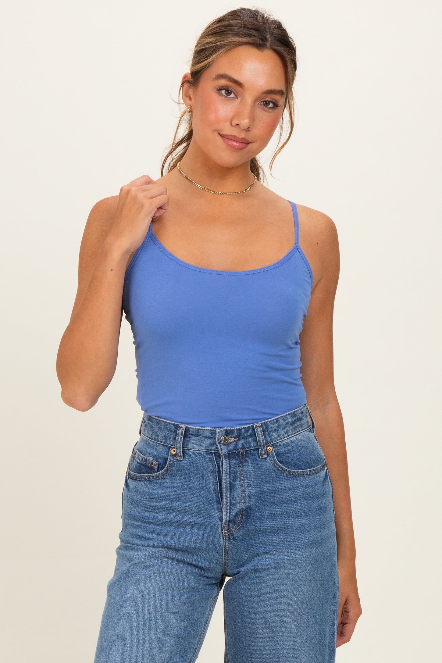 woman wearing a blue fitted camisole top and high-waisted denim jeans, standing with one hand on her shoulder