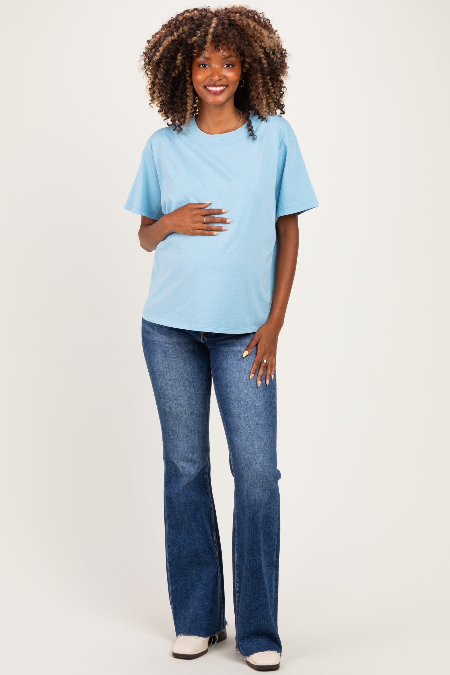 woman wearing a light blue maternity t-shirt and blue flared jeans, gently holding her baby bump