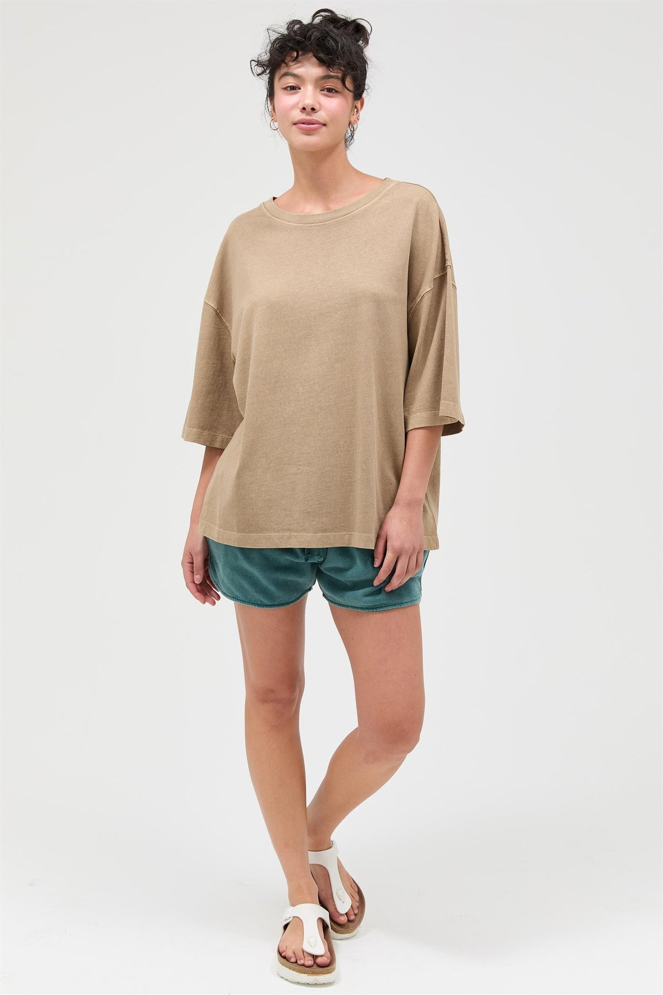 woman wearing a brown oversized short sleeve top and green shorts, standing with one foot slightly forward