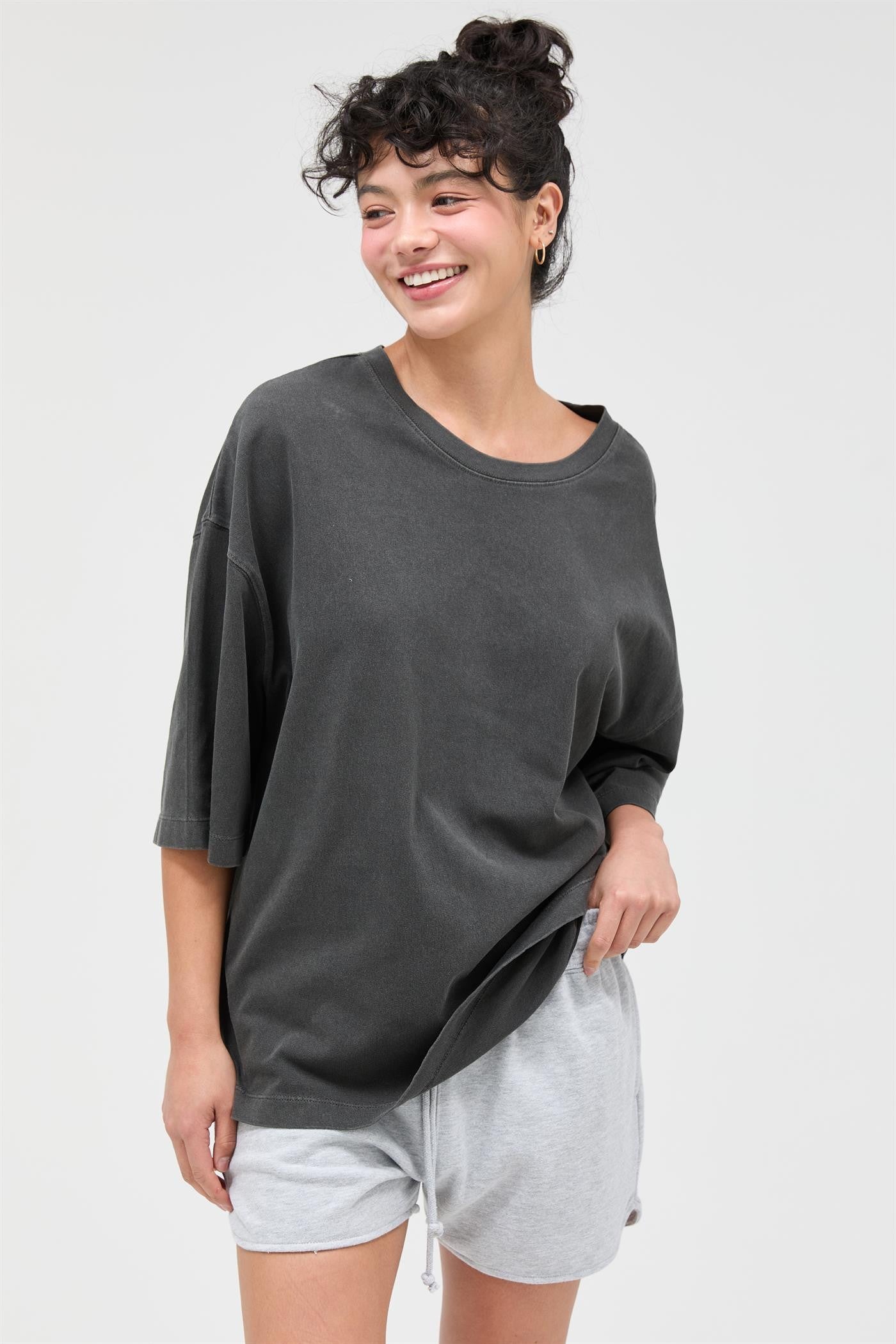 woman wearing an oversized dark gray t-shirt and light gray shorts, smiling and posing casually