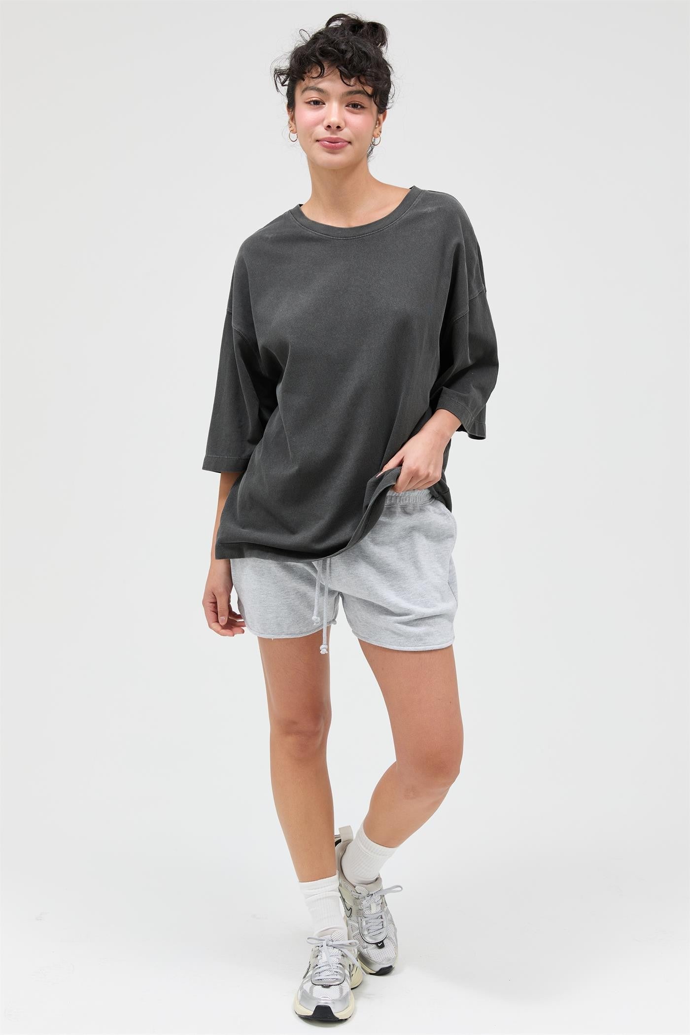 woman wearing an oversized dark gray t-shirt and light gray shorts, standing with one hand in her pocket