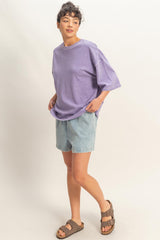 woman wearing a lavender oversized t-shirt and denim shorts, standing with hands in pockets and wearing brown sandals