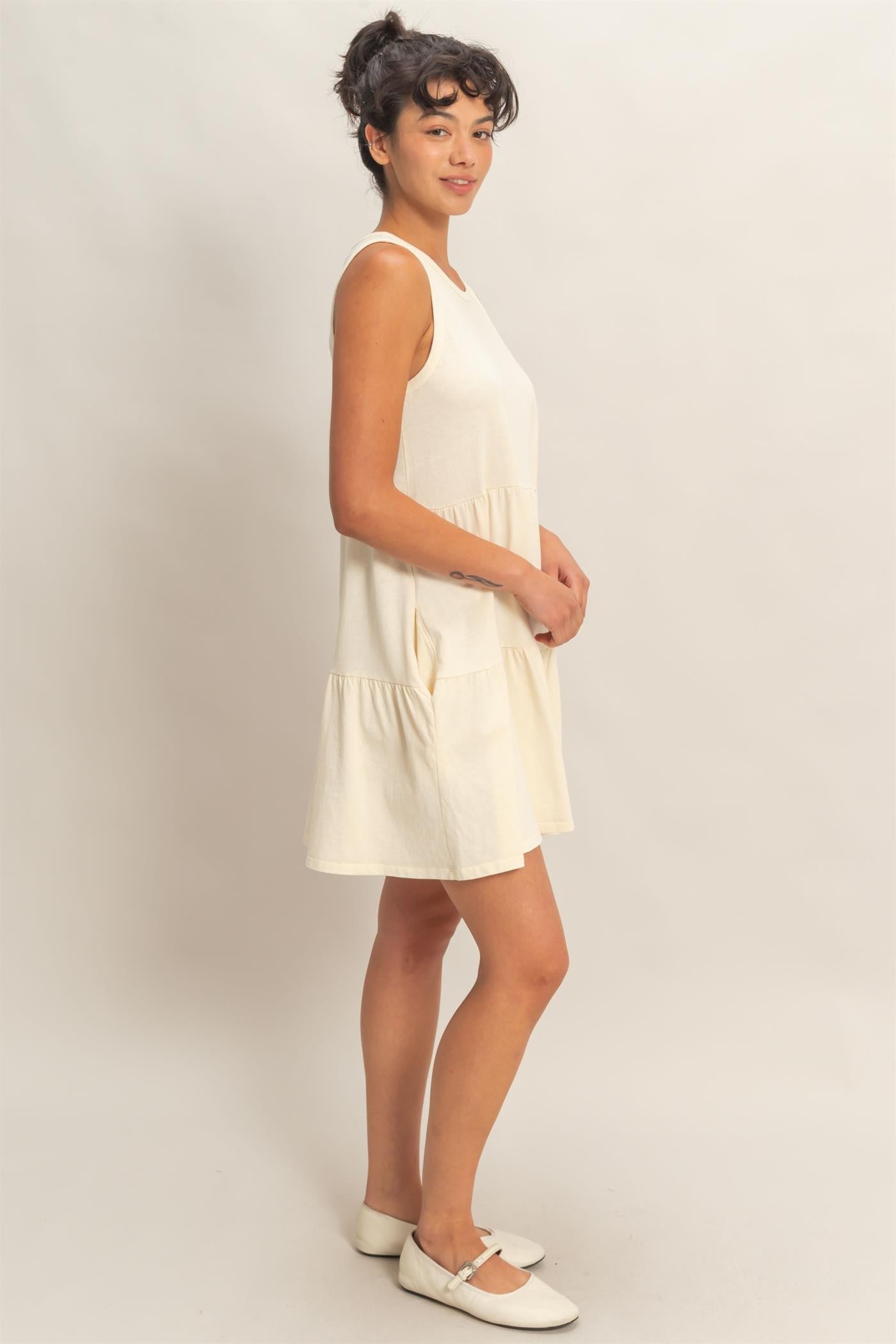 woman wearing a cream sleeveless sundress with a tiered design and pockets, standing against a neutral backdrop