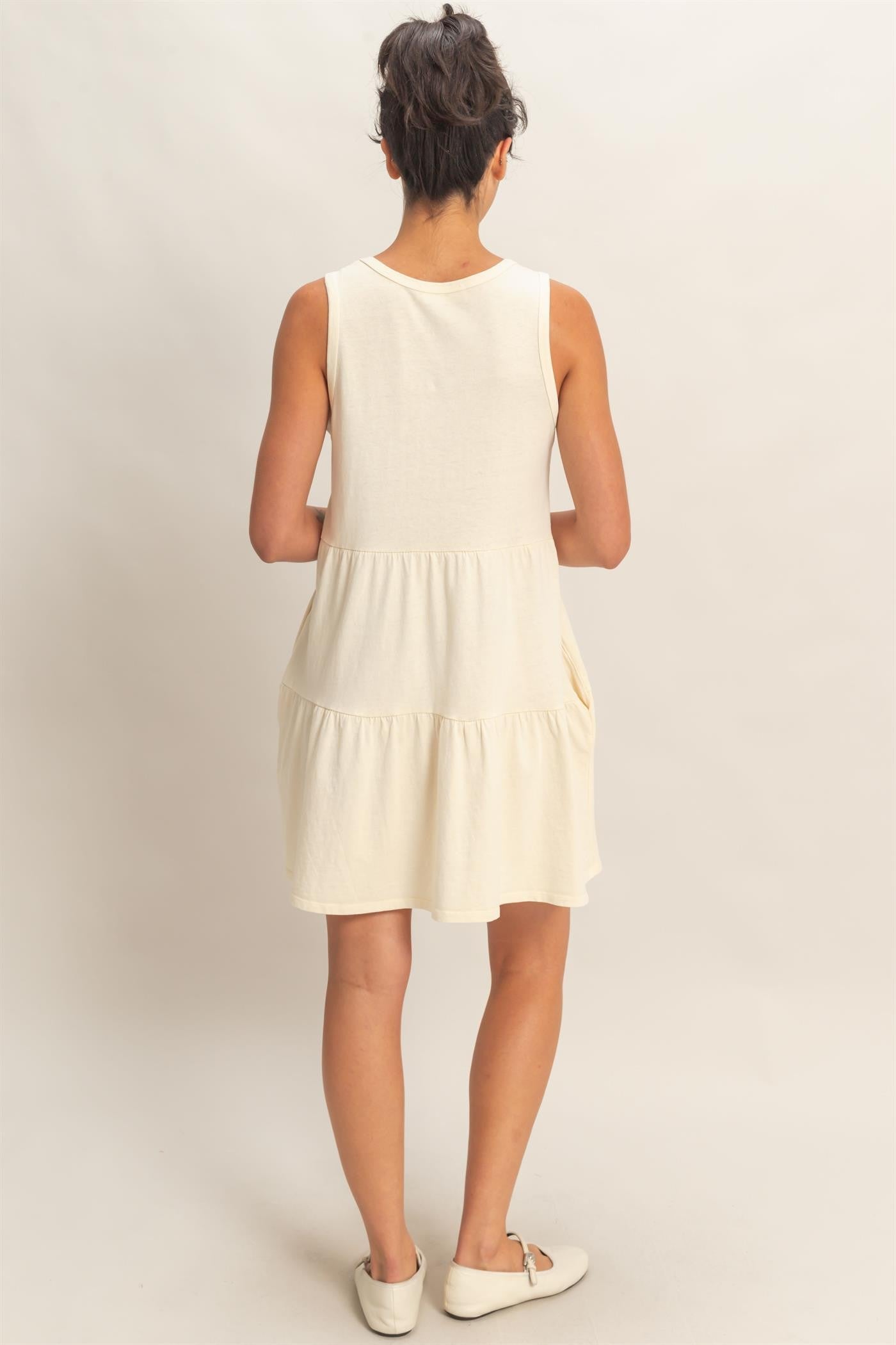 woman wearing a sleeveless cream tiered dress, showing the back view with a relaxed fit and a flared hem