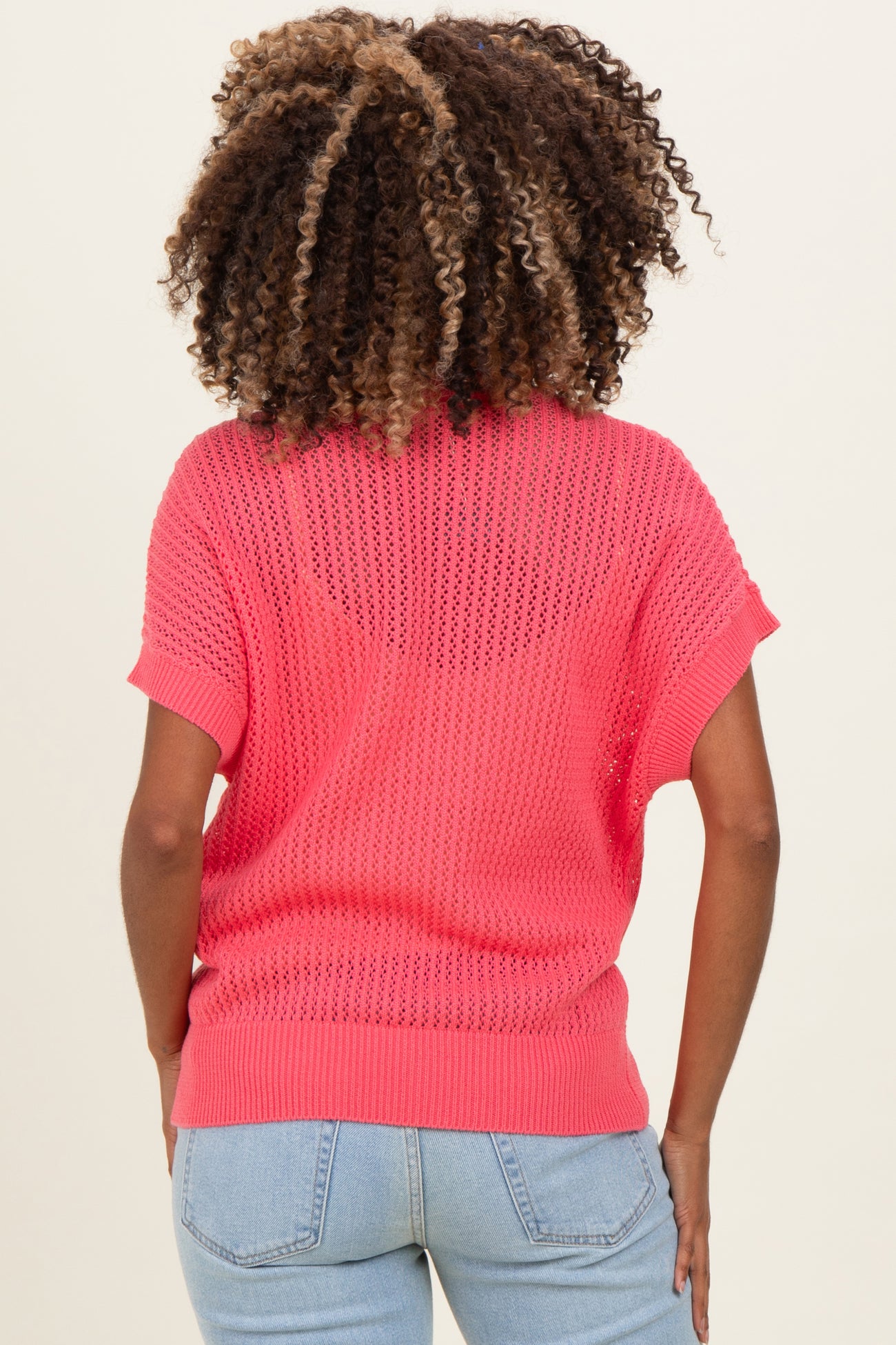 woman wearing a pink open-knit maternity sweater, seen from the back, styled with light blue jeans
