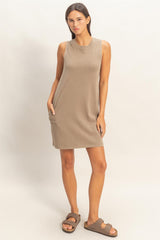 woman wearing a taupe sleeveless dress with side pockets and brown slide sandals