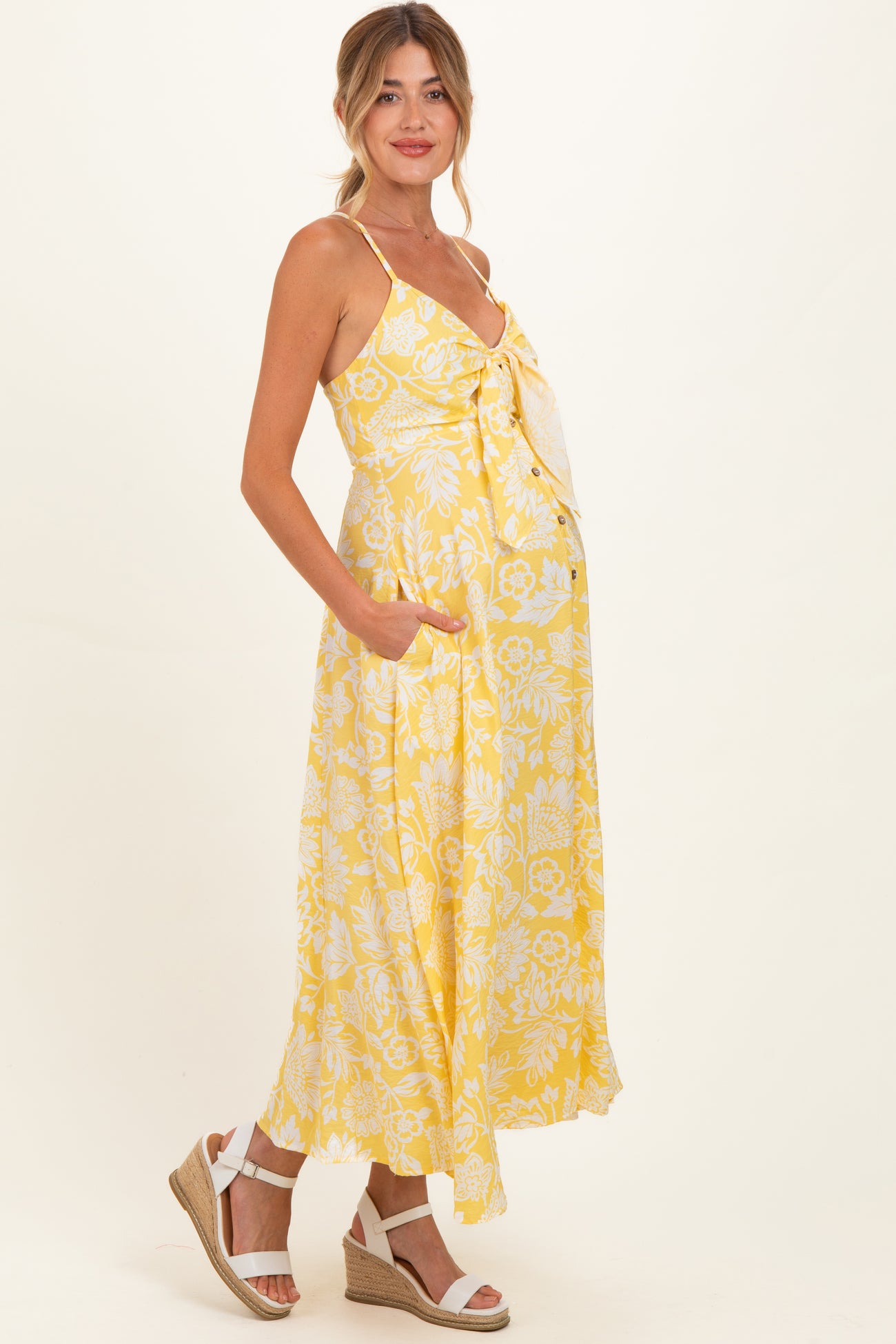 woman wearing a yellow floral maternity maxi dress with adjustable straps and pockets, showcasing her baby bump