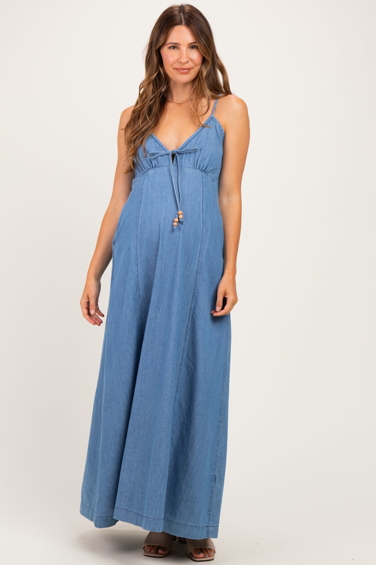 woman wearing a blue denim maternity maxi dress with a tie front and spaghetti straps, showcasing her baby bump