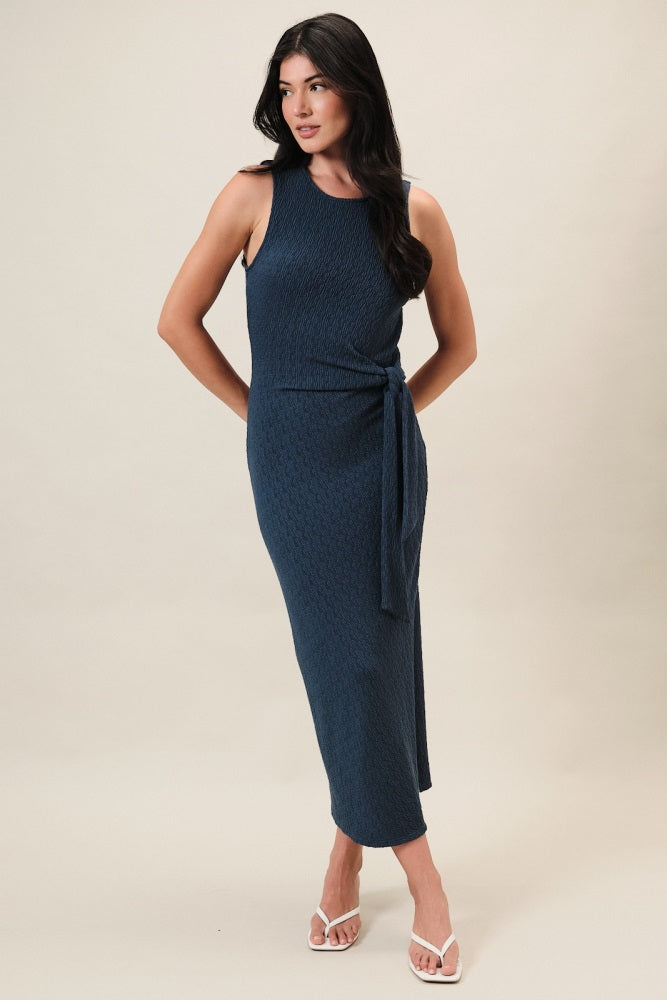 woman wearing a dark blue sleeveless maxi dress with a side tie detail and a fitted silhouette
