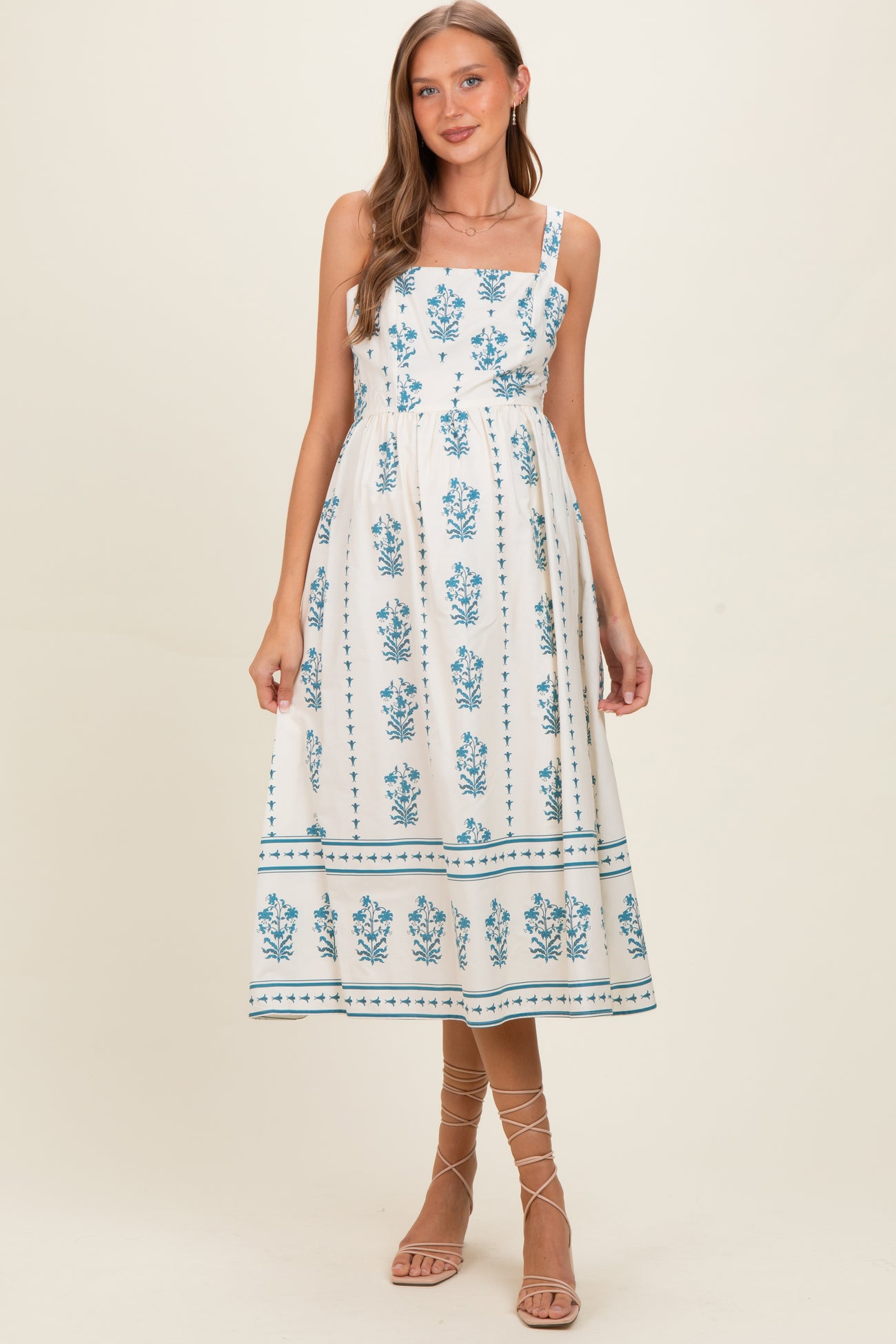 woman wearing a white maternity dress with blue floral print and spaghetti straps, posing without a visible baby bump