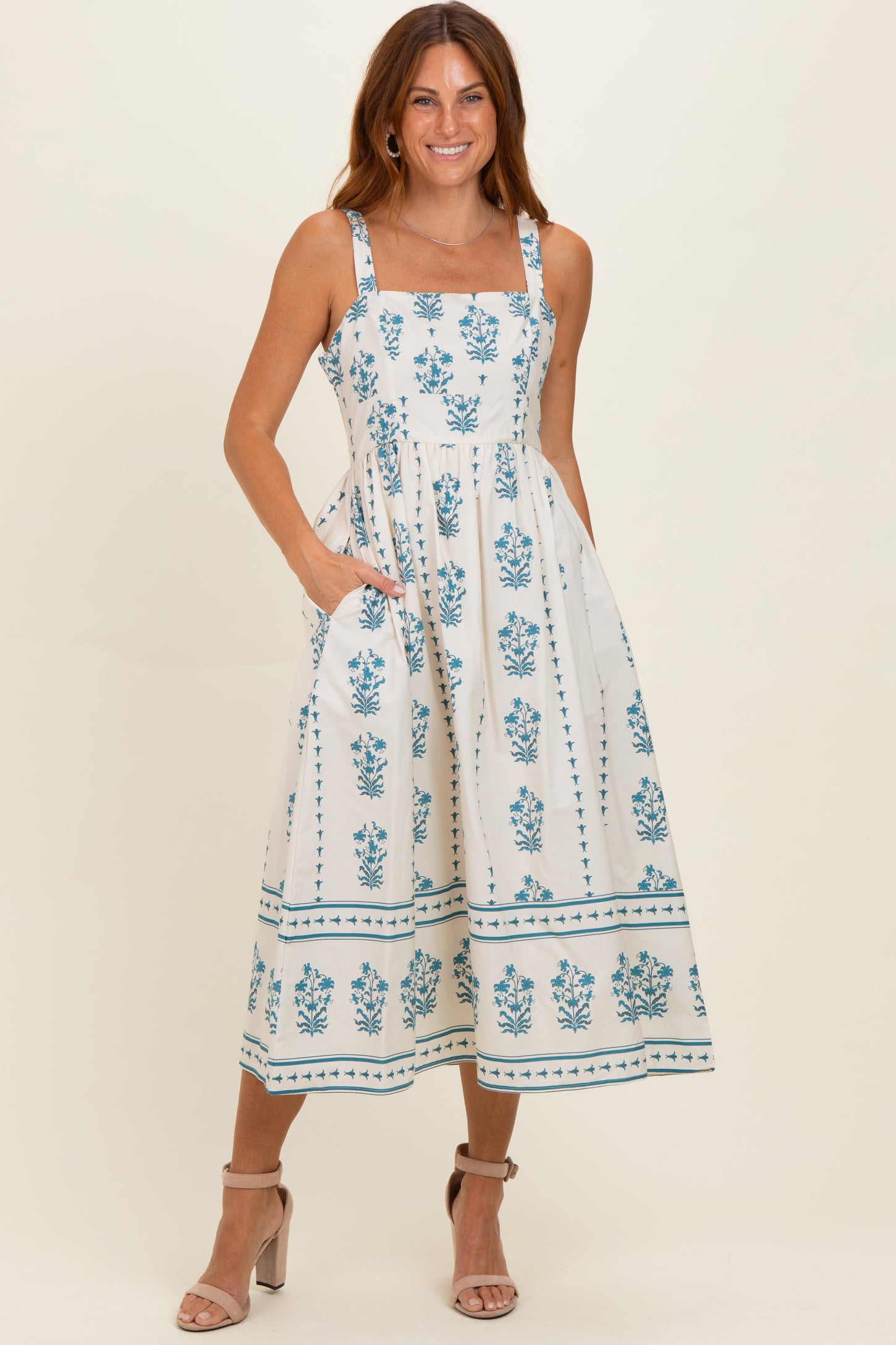 woman wearing a white midi dress with blue floral patterns and square neckline, posing with hands in pockets