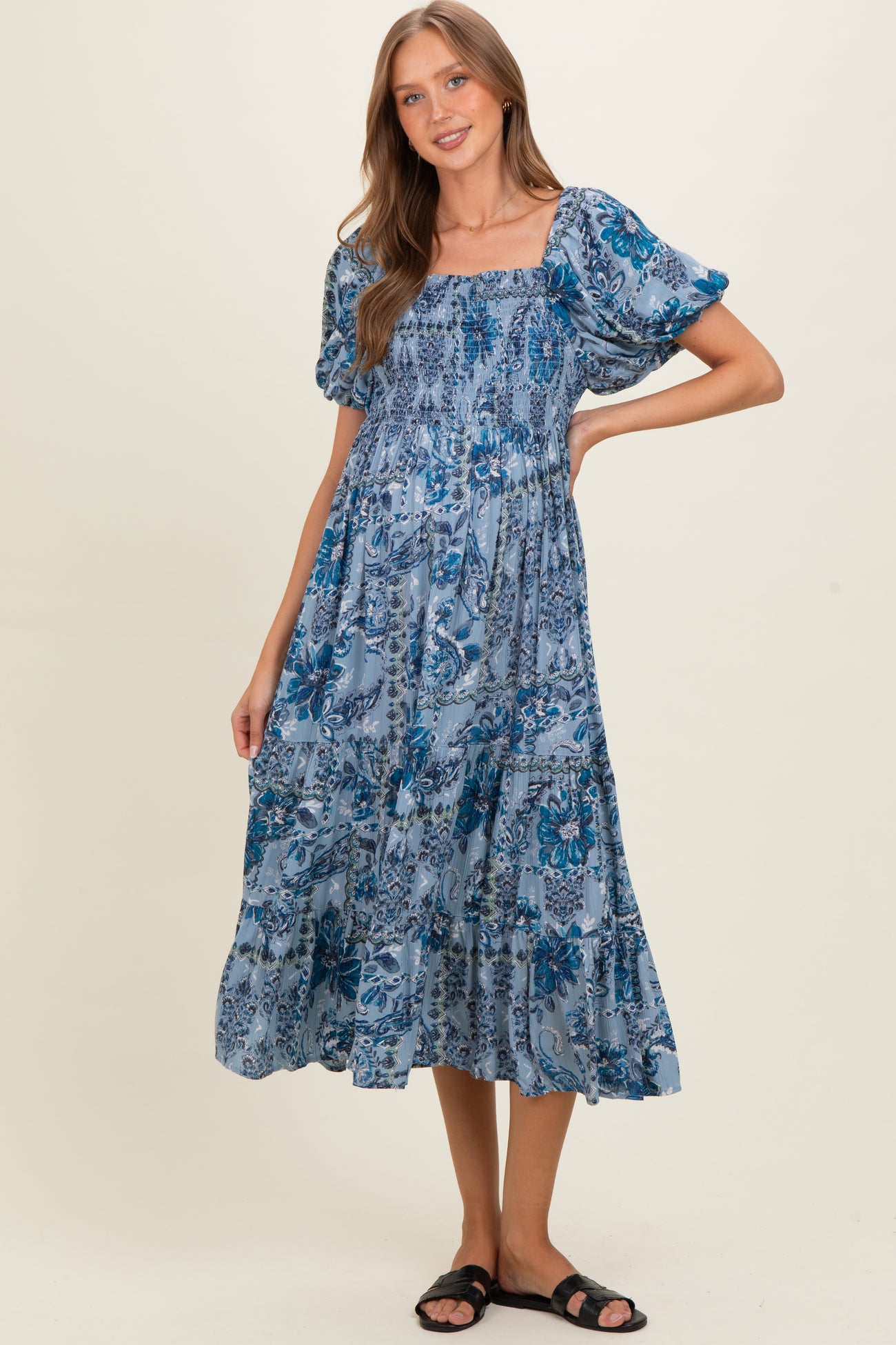 woman wearing a blue floral print maternity dress with puff sleeves and a flowing skirt, smiling with one hand on her hip