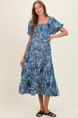 woman wearing a blue floral print maternity dress with puff sleeves and a flowing skirt, smiling with one hand on her hip