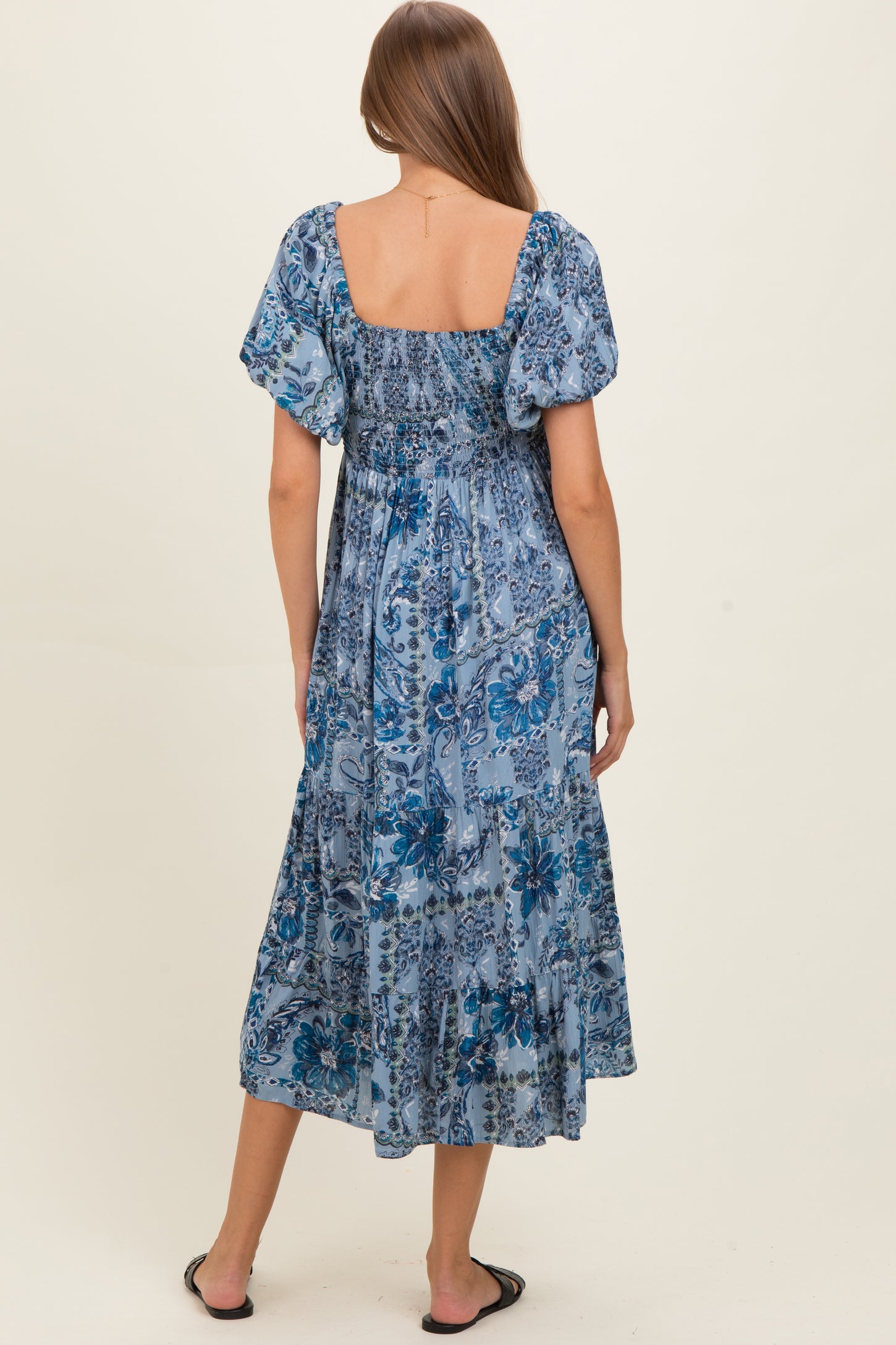 woman wearing a blue floral maternity maxi dress with puff sleeves and a smocked bodice, showing her baby bump from the back