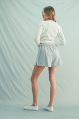 woman wearing a white cropped sweater with floral patterns and gray flared shorts, standing with her back to the camera