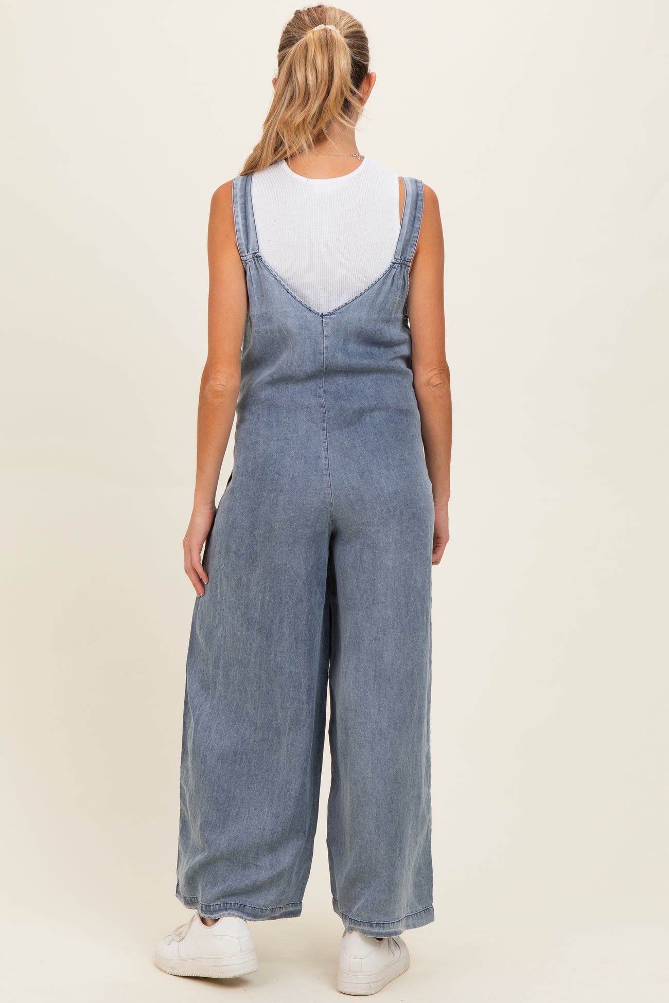 woman wearing a light blue maternity overall jumpsuit with wide leg style and a white tank top underneath, viewed from the back