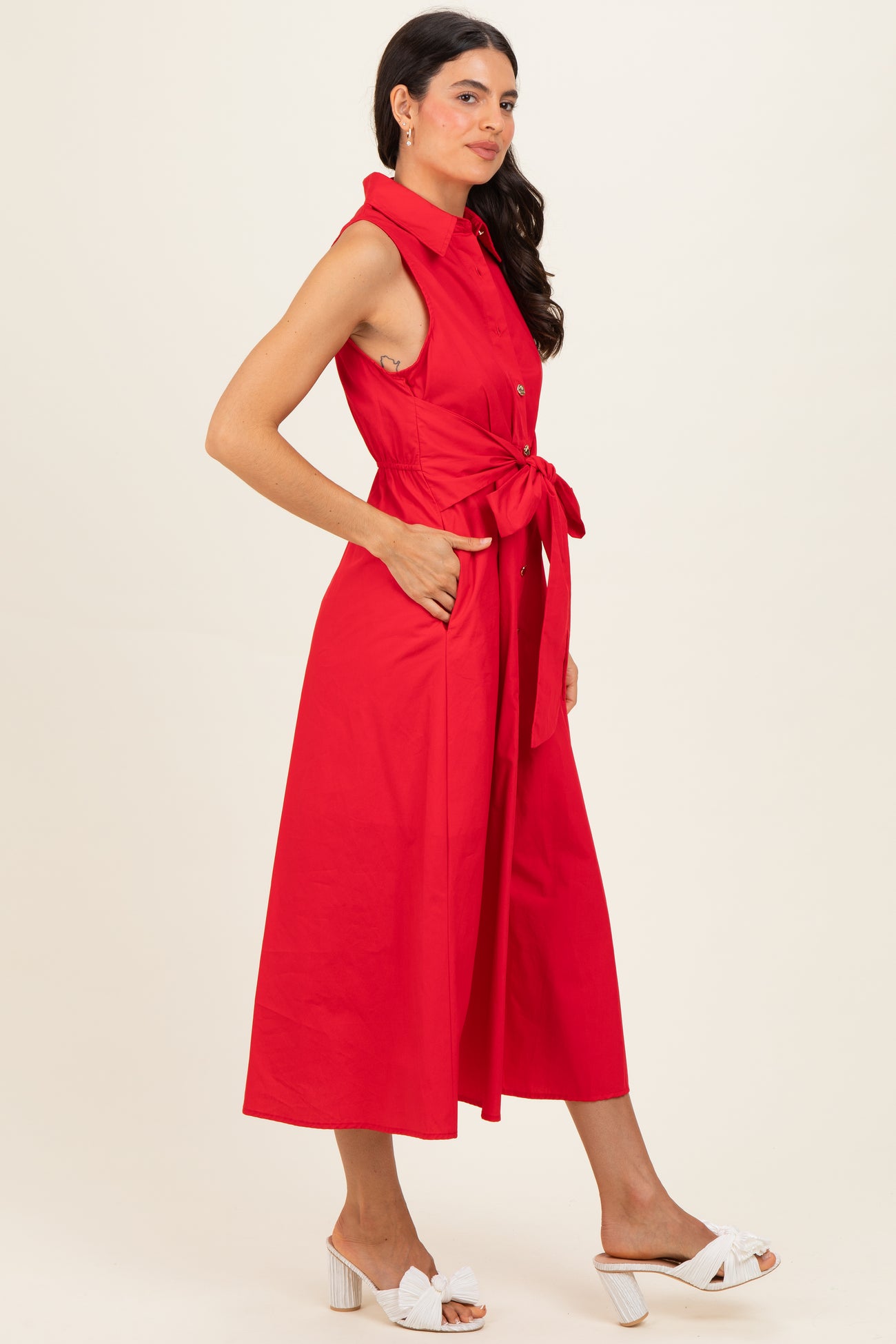 woman wearing a sleeveless red midi dress with a front tie and pockets, styled with white heeled sandals