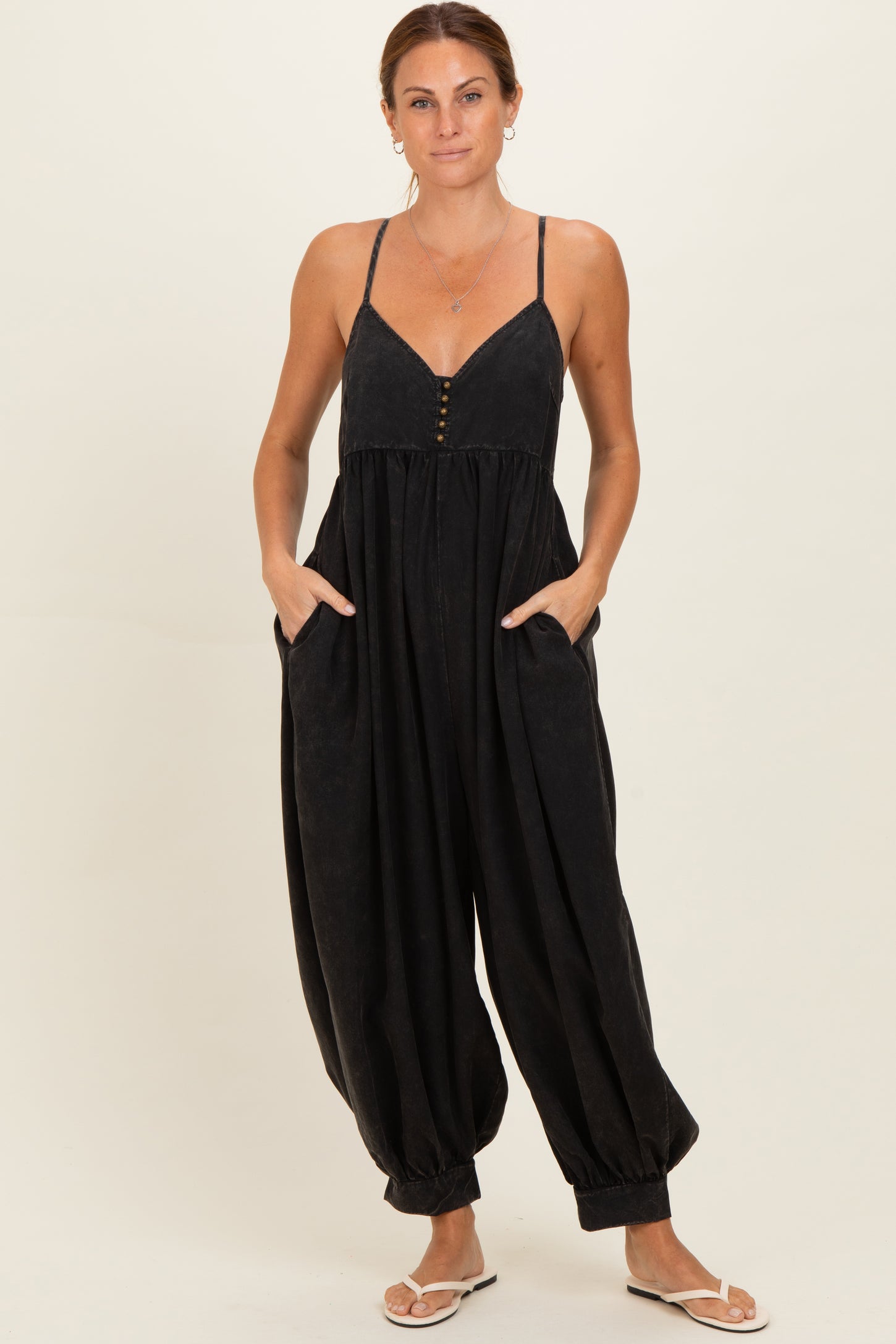 woman wearing a black spaghetti strap jumpsuit with button details and pockets, styled with flat sandals