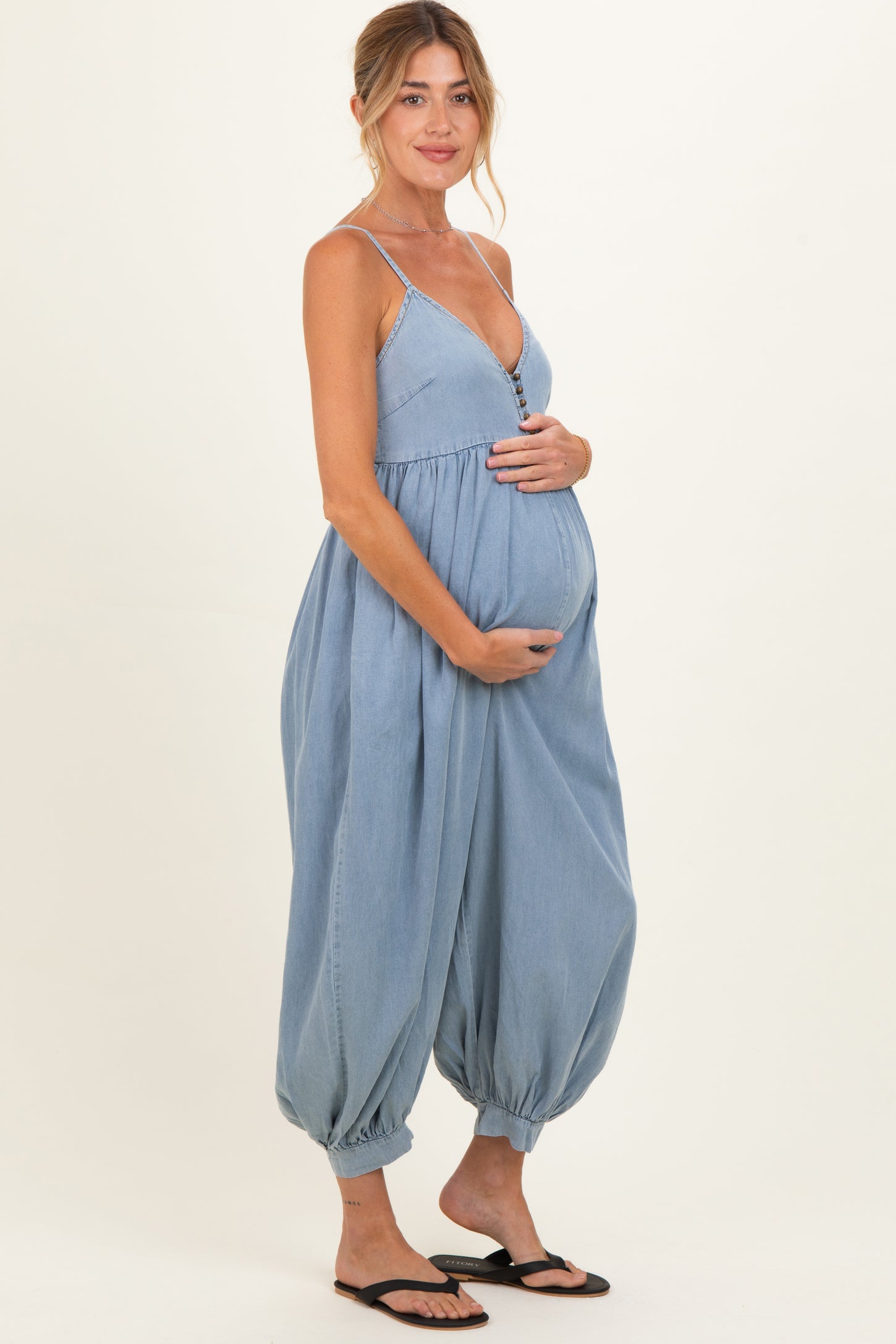 woman wearing a light blue maternity jumpsuit with button details and holding her baby bump