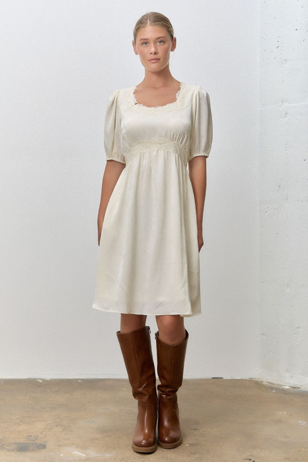 woman wearing a cream short puff sleeve dress with a square neckline and holding brown knee-high boots