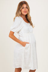 woman wearing a white maternity dress with puff sleeves, floral patterns, and pockets, showing her baby bump