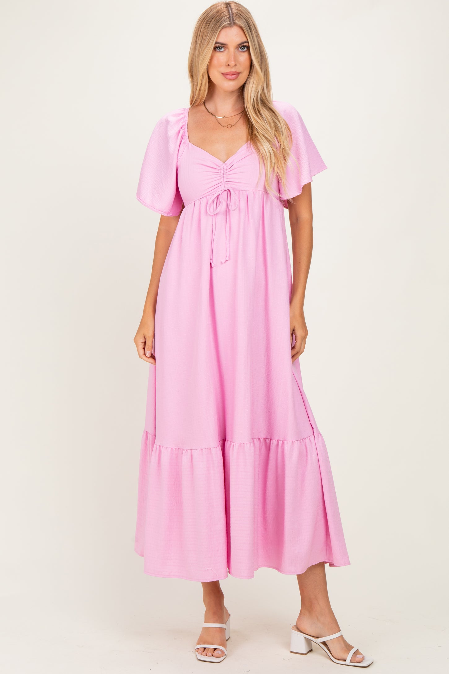 woman wearing a pink maxi dress with puff sleeves and a tie detail at the bust, standing in a light background.