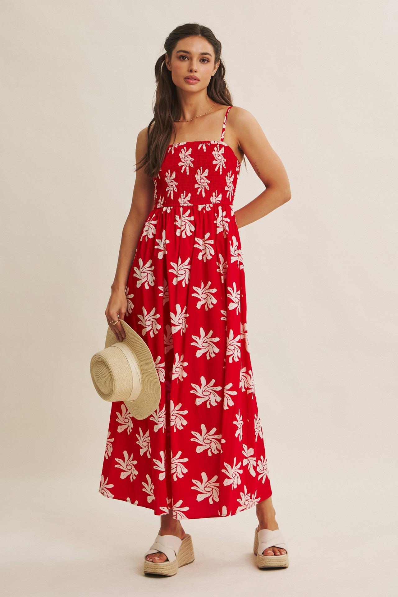 woman wearing a red floral maxi dress with a square neckline and spaghetti straps, holding a straw hat