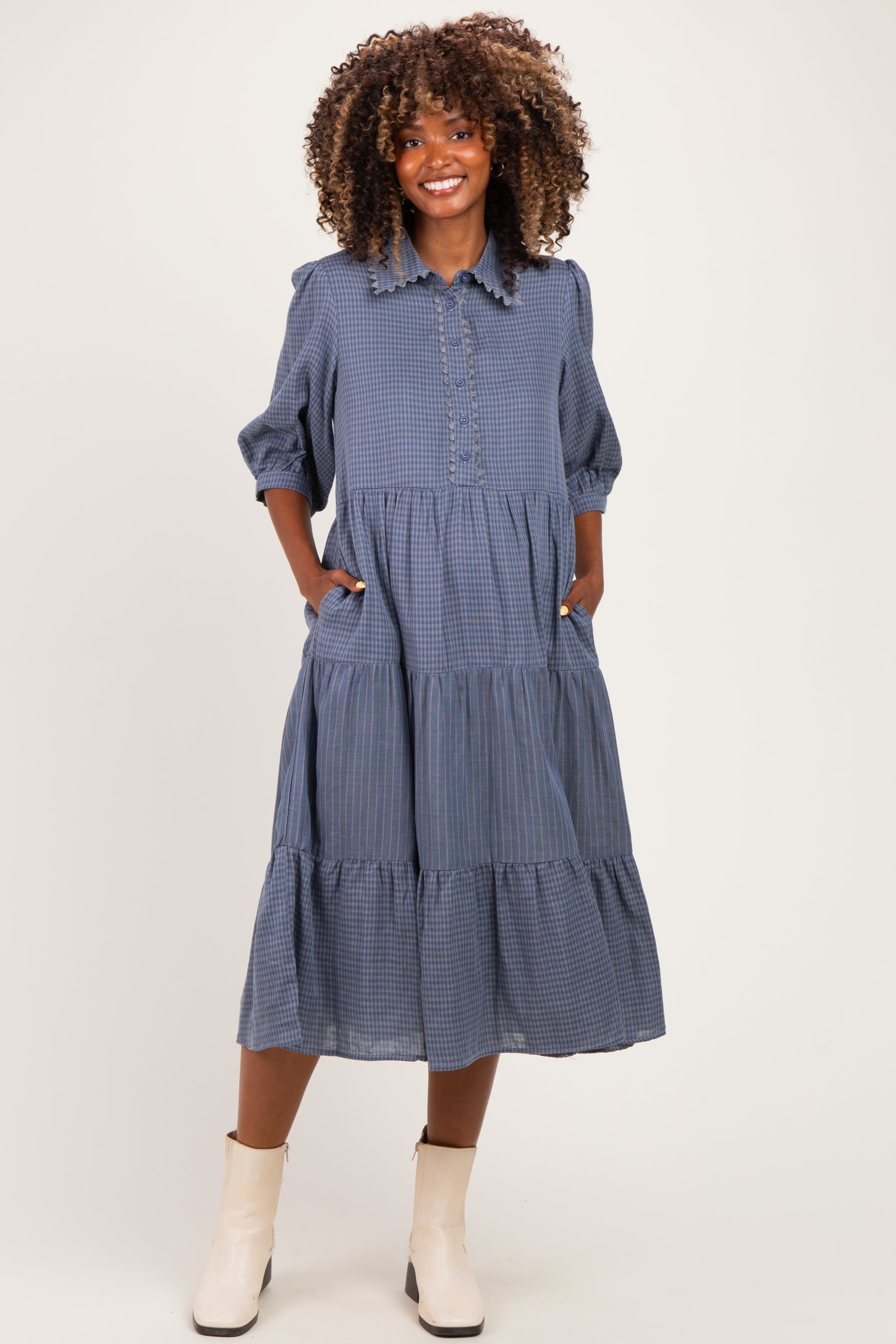 woman wearing a blue gingham maternity dress with a button-up front, three-quarter sleeves, and tiered skirt, styled with ankle boots