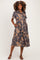 woman wearing a floral maternity dress with short sleeves, a tie waist, and pockets, standing against a plain background