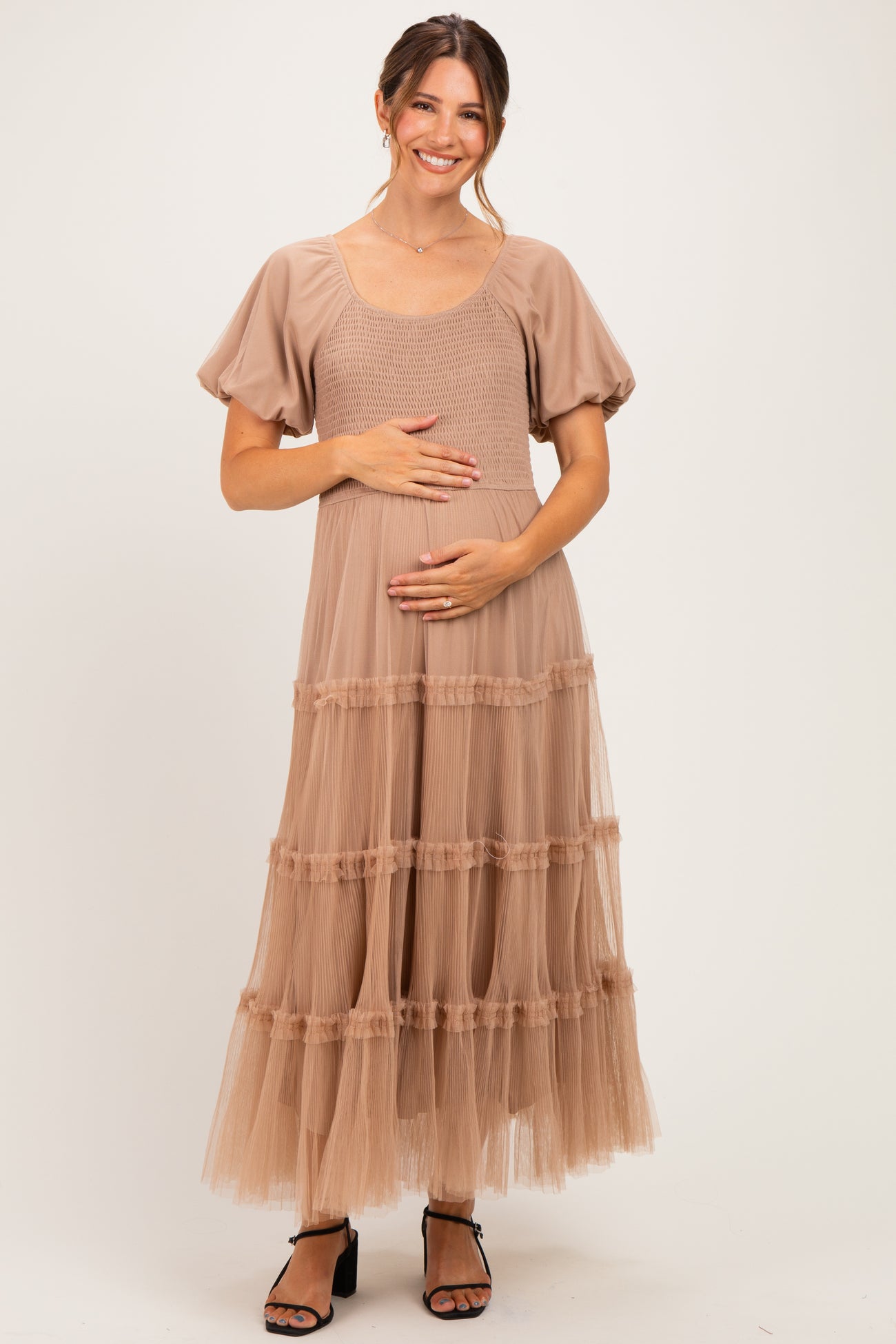 woman wearing a taupe maternity dress with puff sleeves and tiered tulle skirt, holding her baby bump