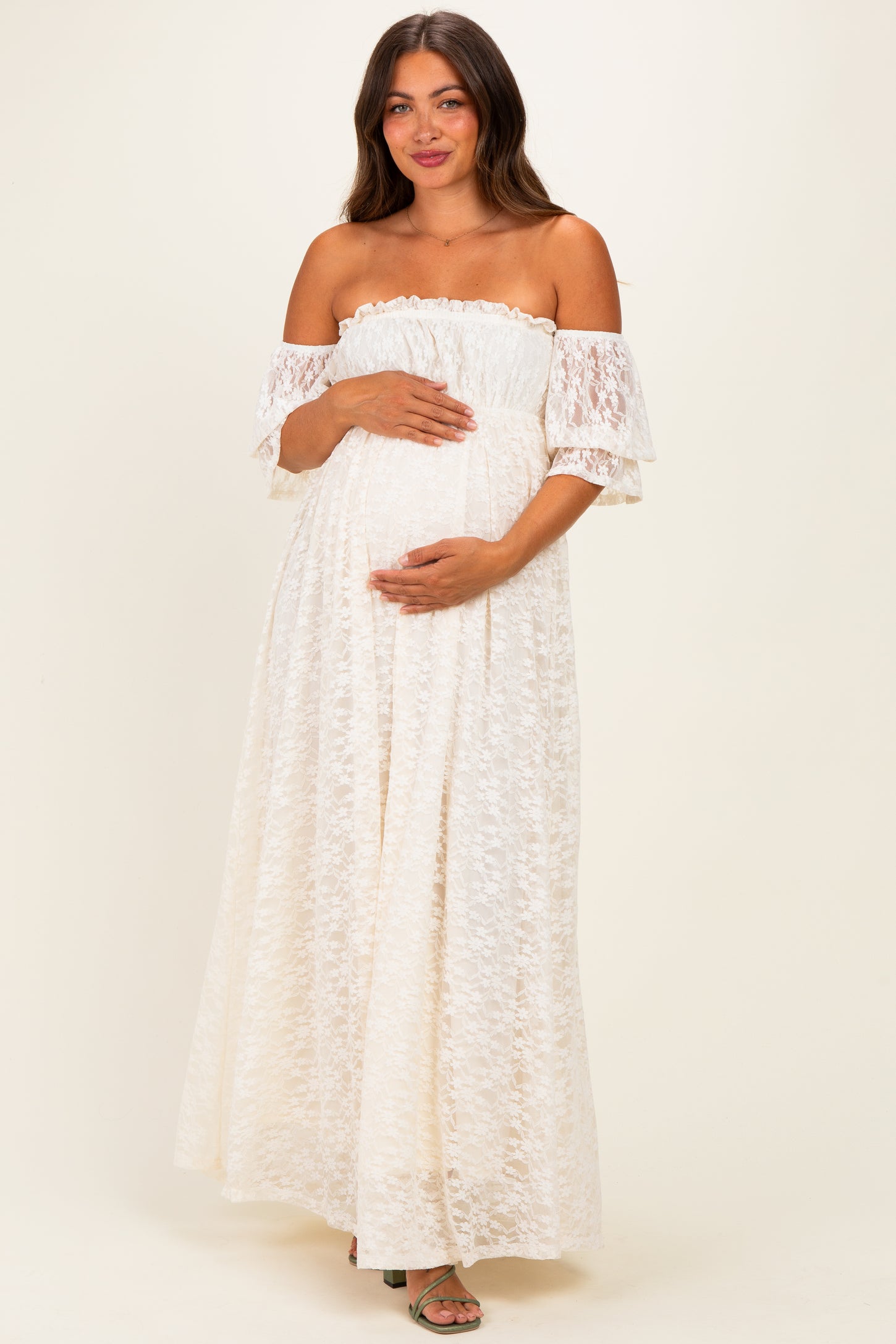 woman wearing a white off-the-shoulder lace maternity dress and holding her baby bump