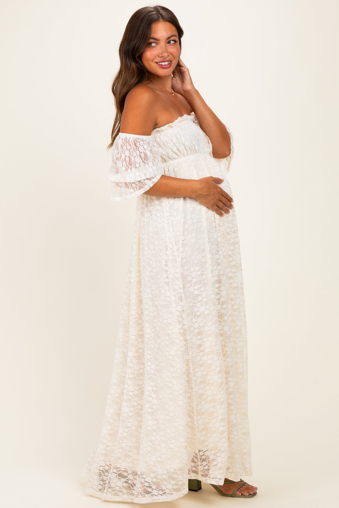 woman wearing a white off-the-shoulder lace maternity dress and holding her baby bump