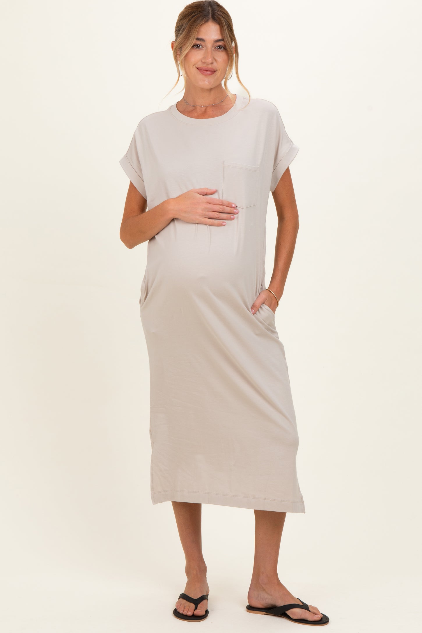woman wearing a light beige maternity dress with short sleeves and side pockets, holding her baby bump