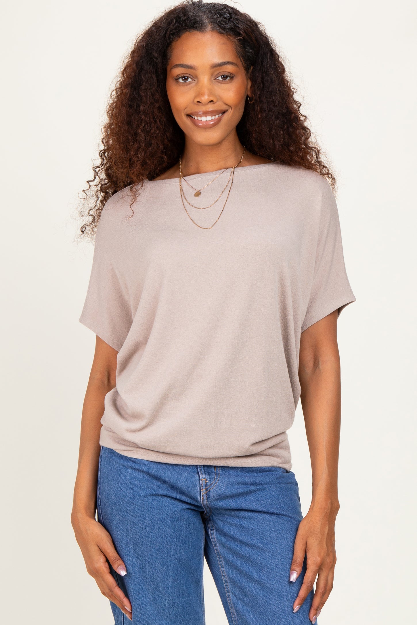 woman wearing a taupe loose-fitting short sleeve top with a round neckline and layered necklaces, paired with denim jeans