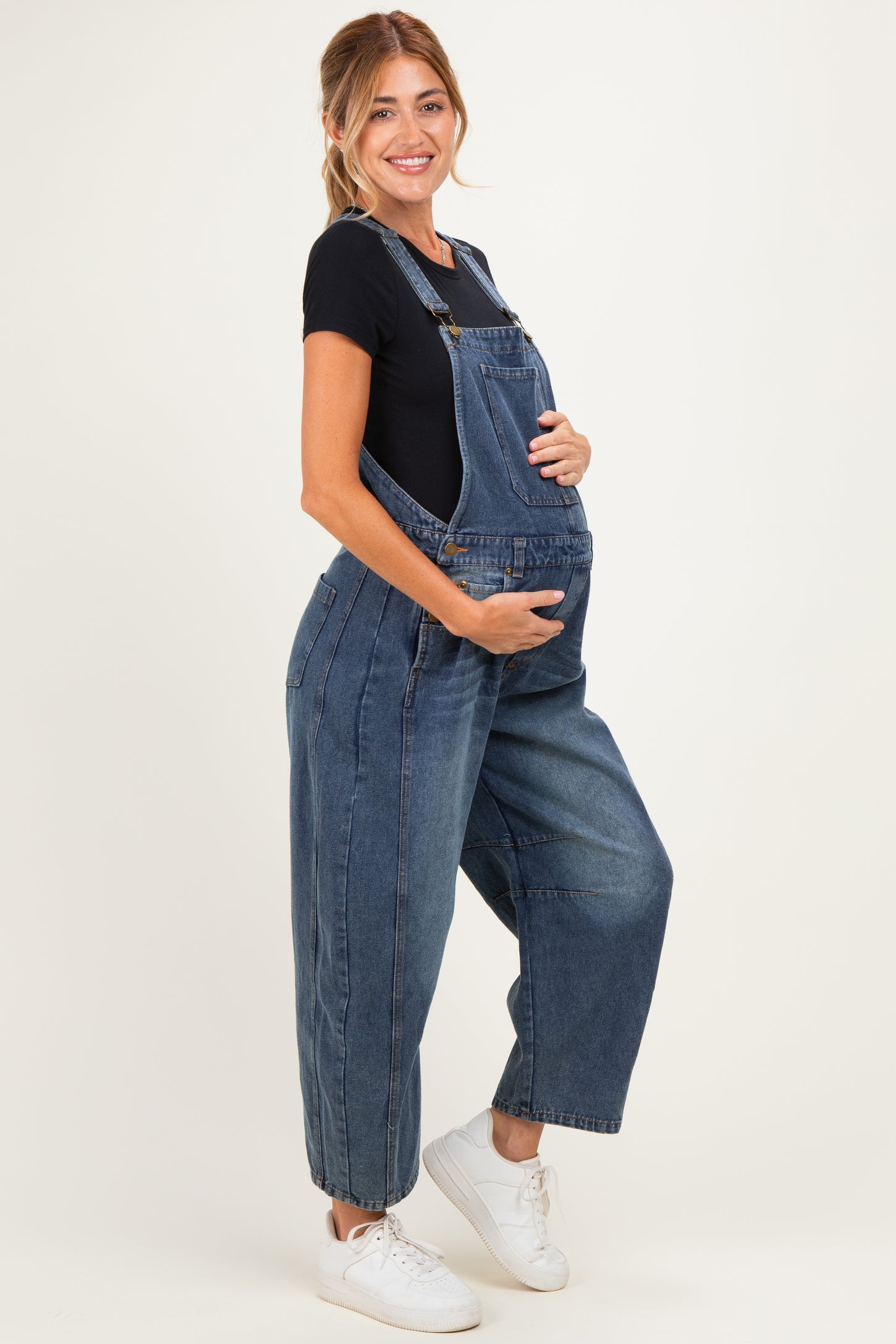 woman wearing a dark blue denim maternity overall, holding her baby bump with a black t-shirt underneath